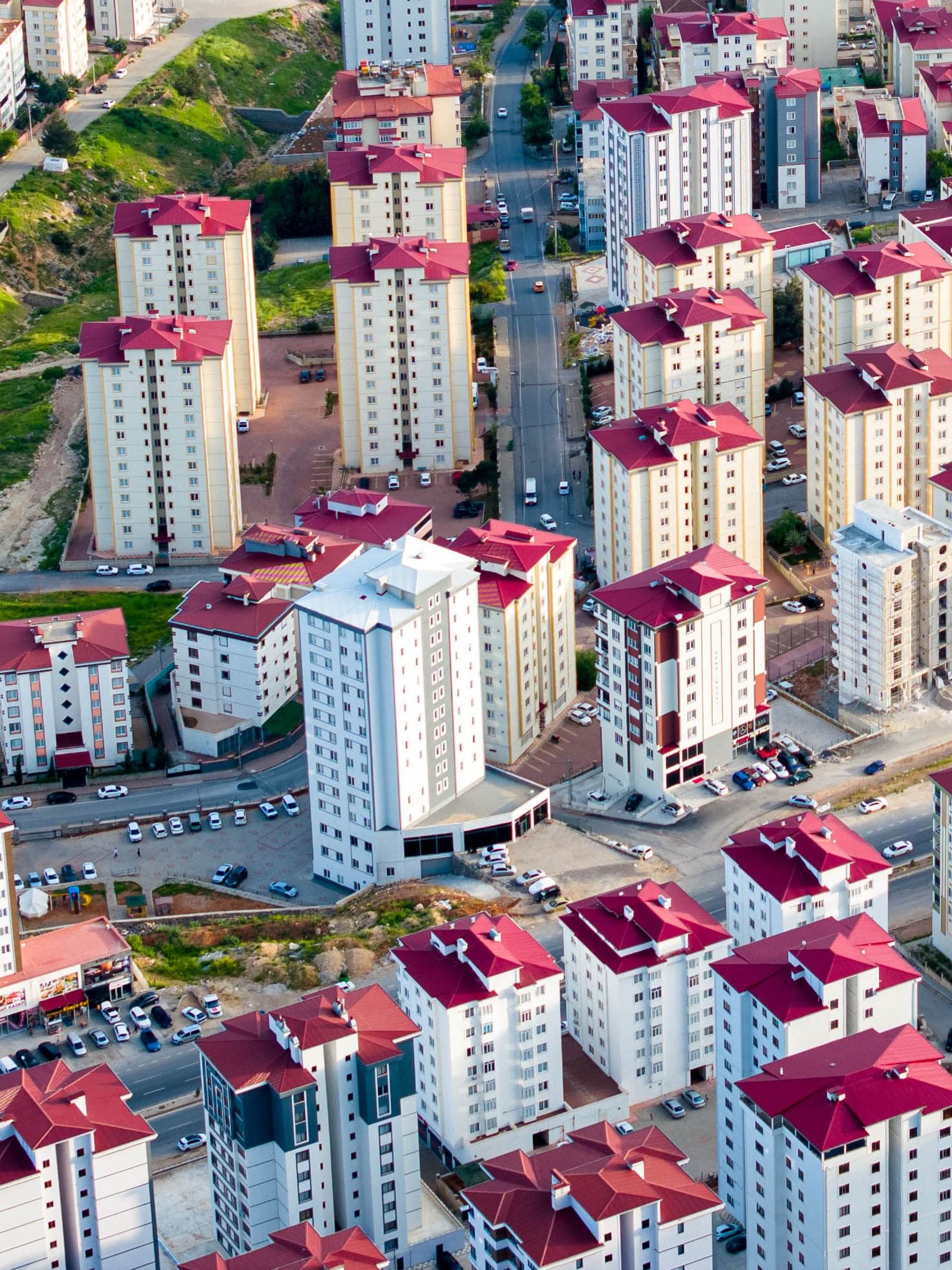 Completed mid-rise residential blocks in Kahramanmaraş seen from height, with the internal roads, parking and planting that serve them and the ground falling away at the edge of the scheme.