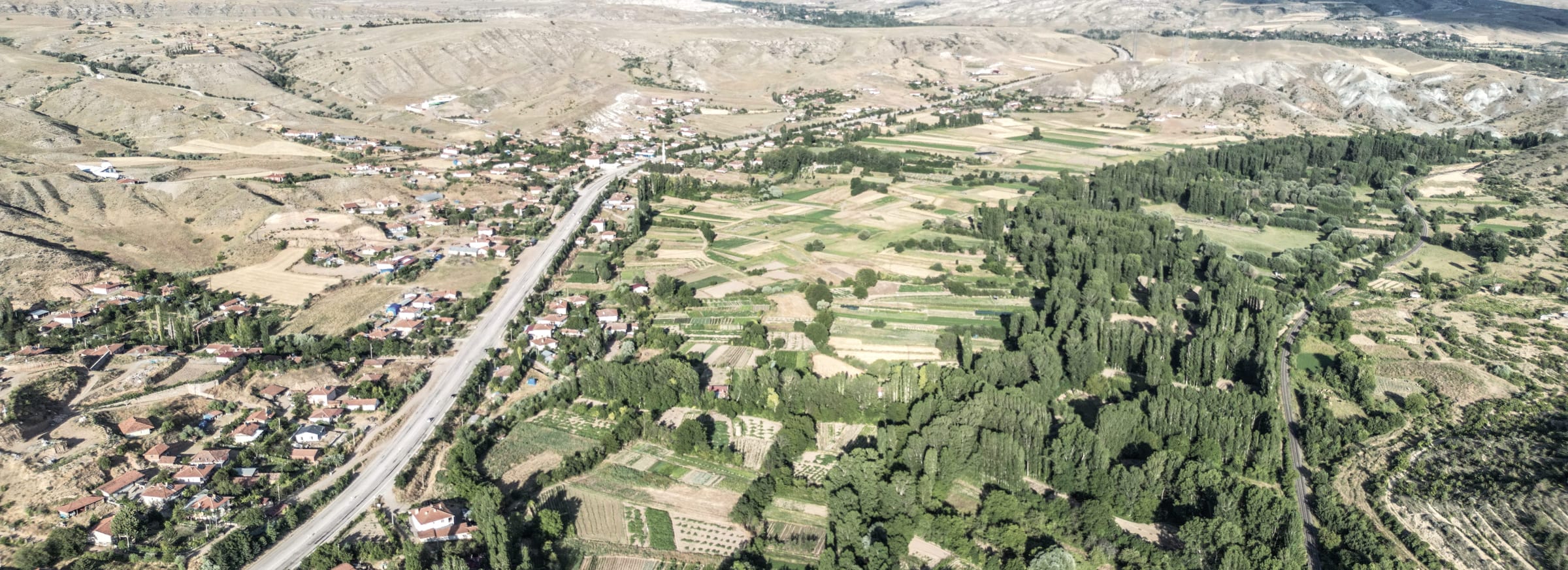 A Çankırı valley read from height: a road running the length of the frame with a village strung along it, irrigated parcels in many hands to one side, a tree belt following the water, and dry hillside closing both edges.
